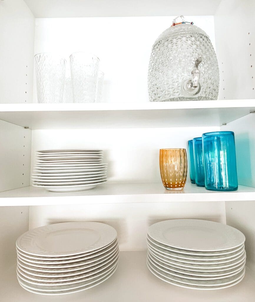 Stacked white plates and colorful glasses on white shelves.