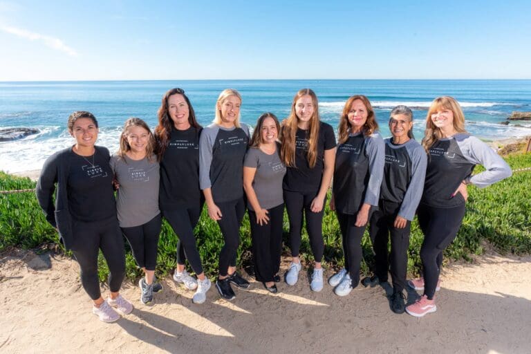 A group of nine women standing by the ocean, smiling and dressed in casual gray and black outfits.