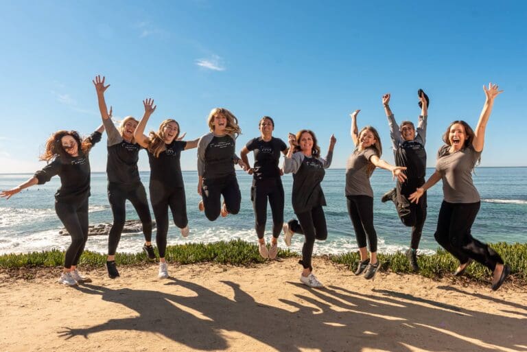 A group of seven people jumping joyfully on a sunny day by the sea.