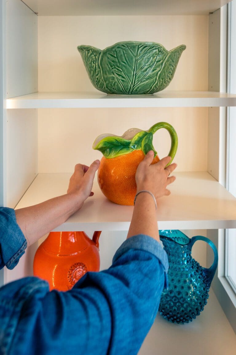 Organizer placing an orange pitcher on a styled open kitchen shelf with green ceramics