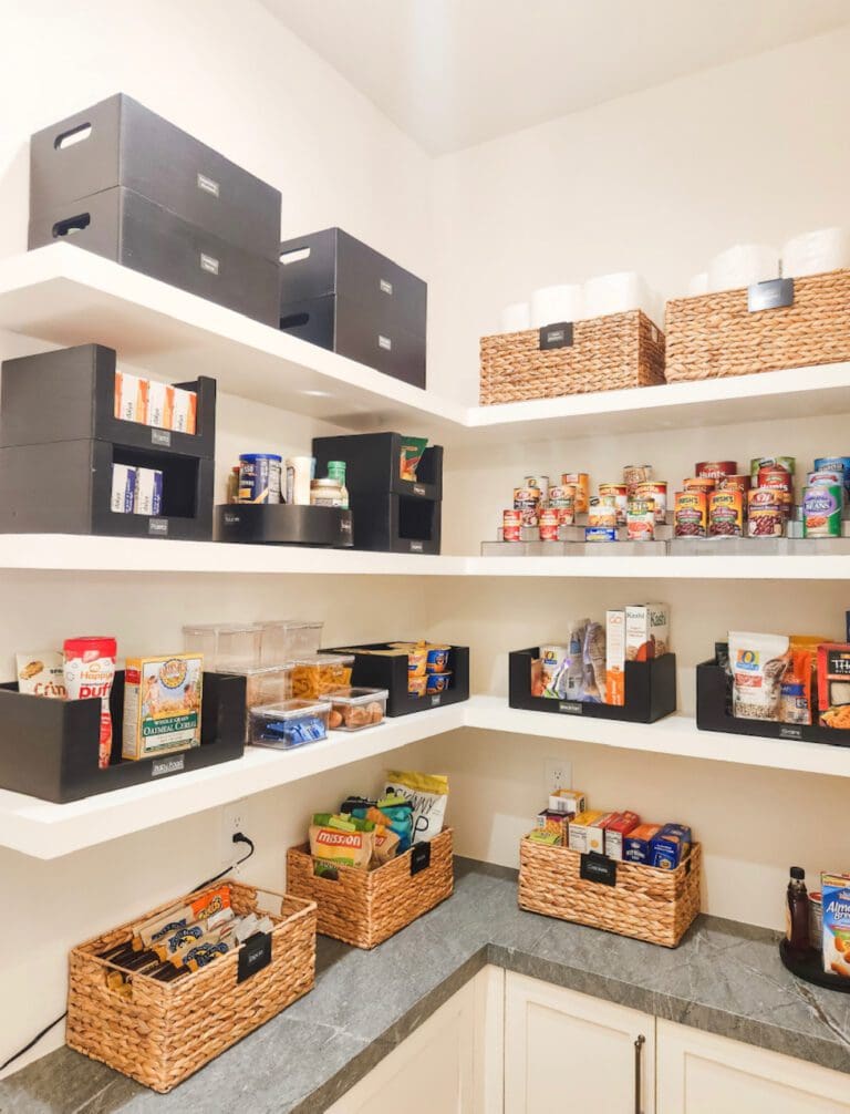 Corner pantry with white shelves, woven baskets and black bins sorting snacks and canned goods