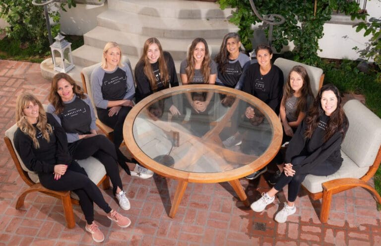Group of people sitting and standing around a round glass table outdoors.