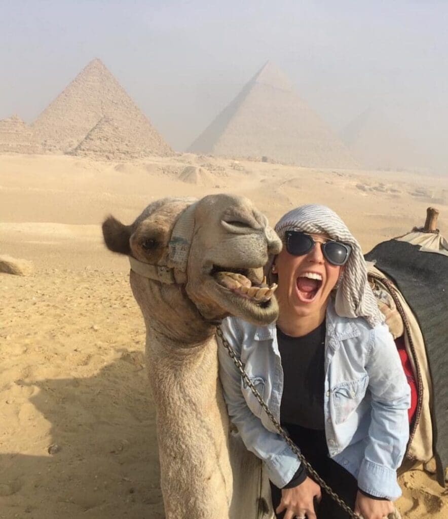 A joyful woman posing with a smiling camel near the pyramids in the desert.