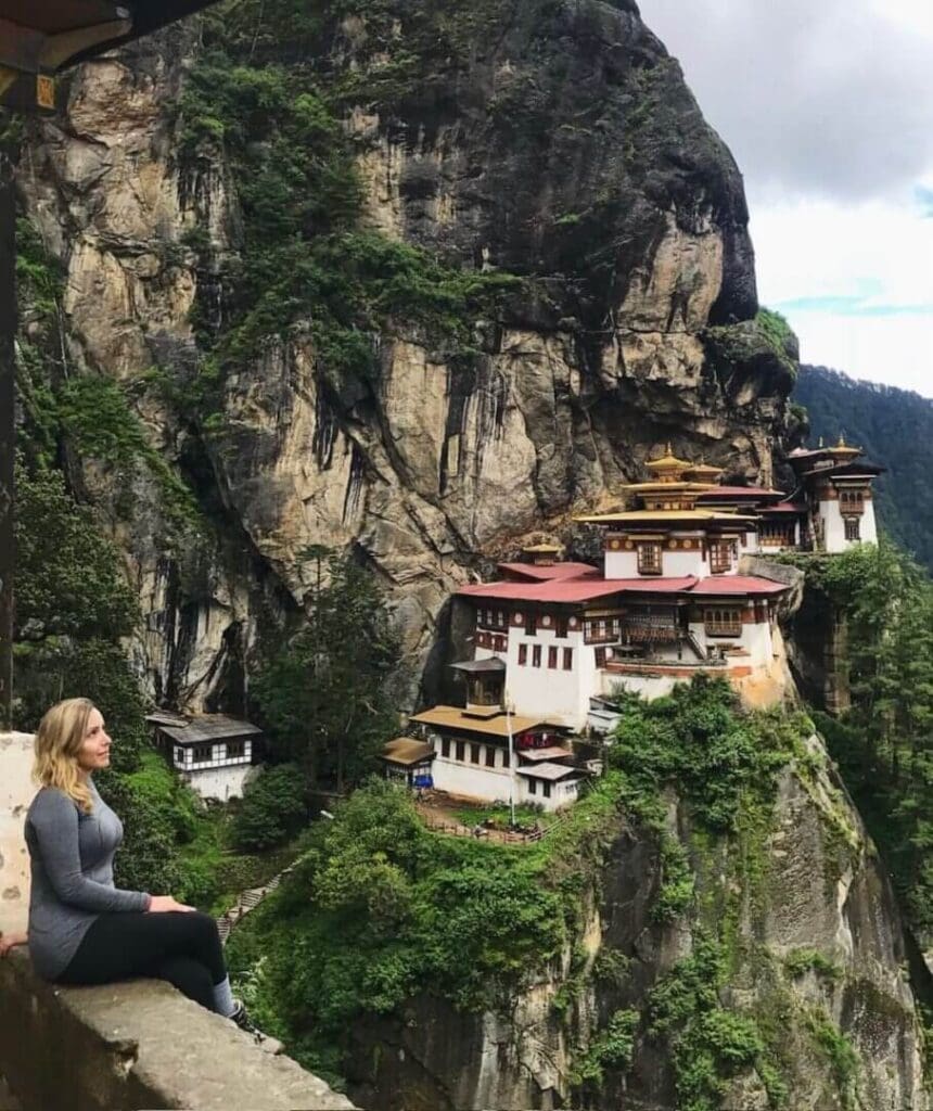 A woman meditating near a cliffside monastery amidst lush greenery.