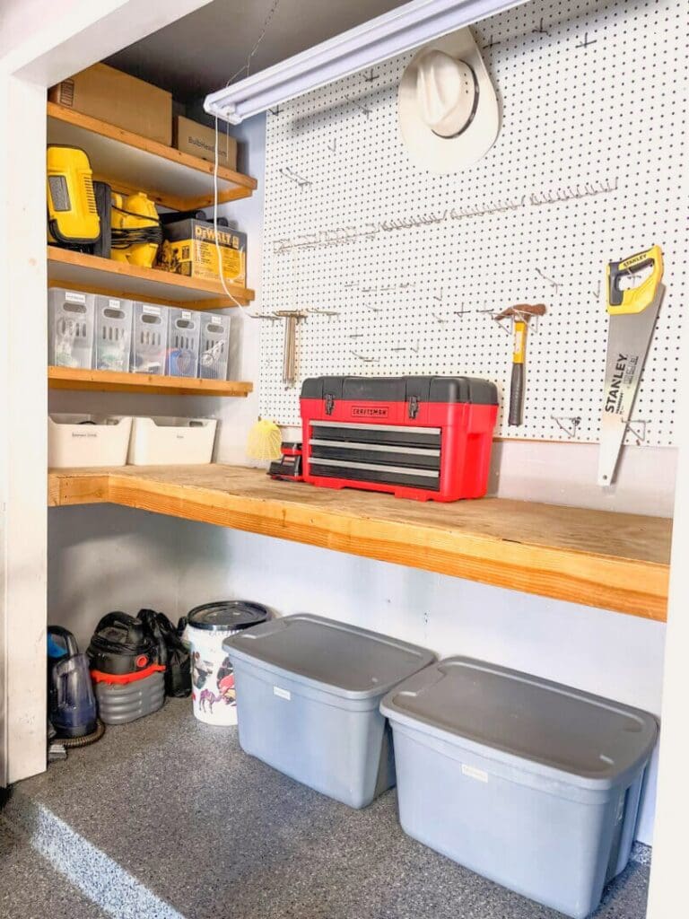 Garage workshop corner with tools on pegboard, bins and a red toolbox on a wood bench