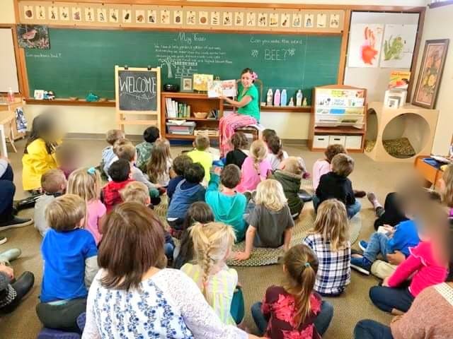 Ryen Toft reading a picture book aloud at the front of a classroom at a school in Orange County while students sit on the rug listening