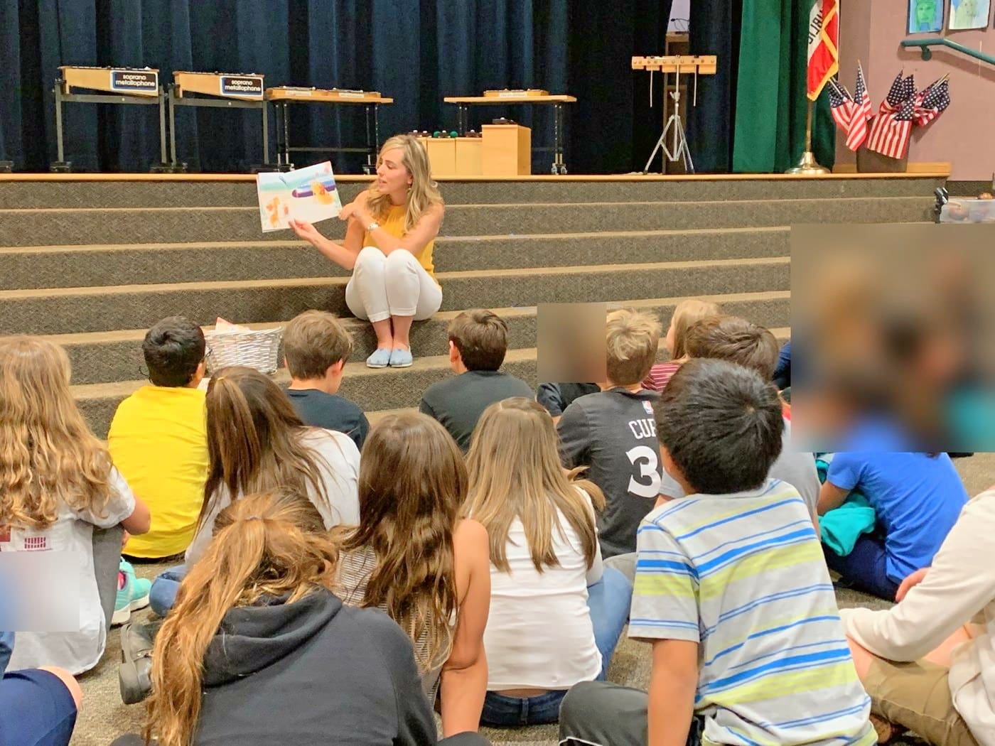 Ryen Toft sitting on the stage steps reading Bee A Dreamer aloud to students at an elementary school in Rancho Santa Fe