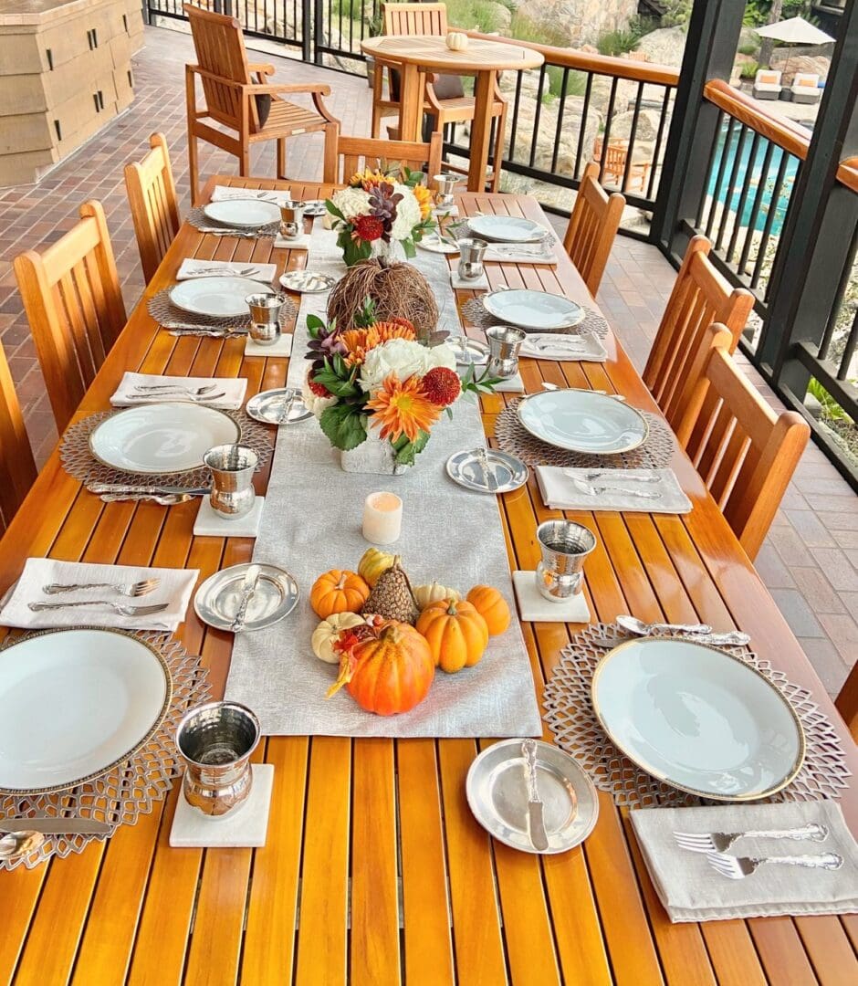Long outdoor patio table set for a fall gathering with white plates, a burlap runner, mini pumpkins and a floral centerpiece, styled by Simply Luxe