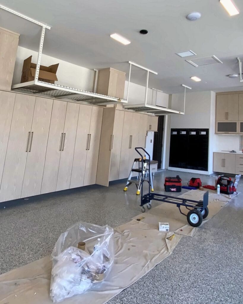 Garage during the California Closets cabinet build-out, with empty overhead racks, a hand truck and drop cloths on the epoxy floor, San Diego