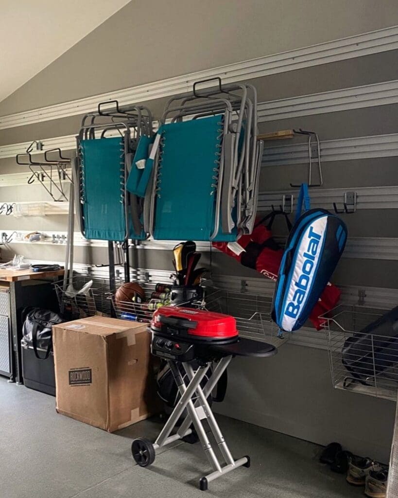 Garage slat wall before organizing, with folding lawn chairs piled on hooks, a tennis bag, a portable grill and a box on the floor