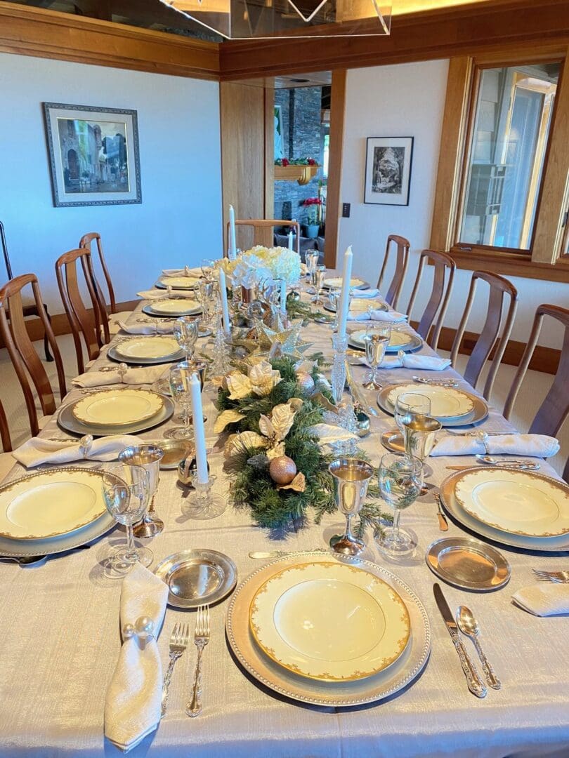 Long dining table set for a holiday dinner with gold-rimmed china, silver goblets, white taper candles and a greenery centerpiece, styled by Simply Luxe