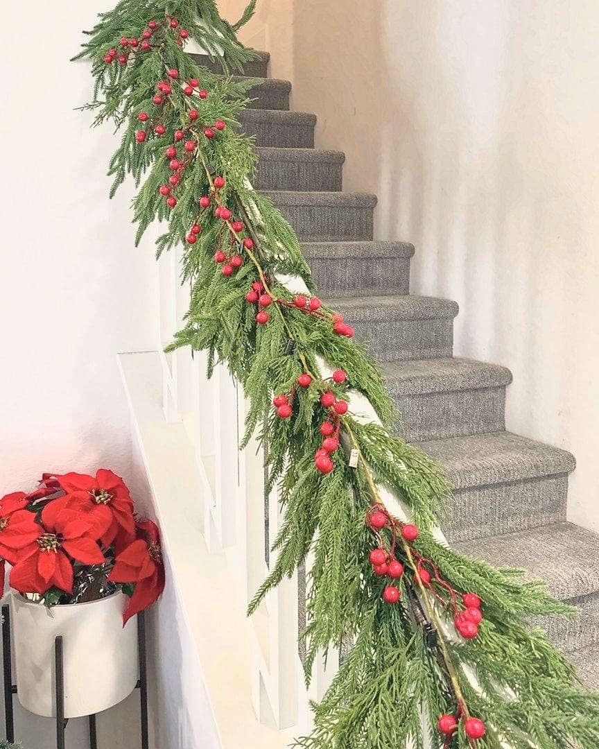 Staircase railing wrapped in fresh greenery garland with red berries and a poinsettia planter, holiday decorating by Simply Luxe