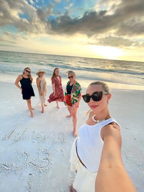 Five women posing happily on a sandy beach during sunset.