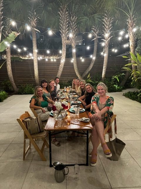 Group enjoying a festive outdoor dinner under string lights and palm trees.