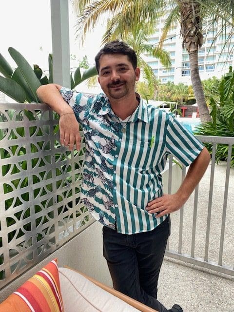 Man in a striped shirt poses casually by a white fence outdoors.