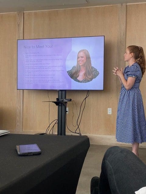 A woman in a blue dress presents next to a screen with text and a portrait.