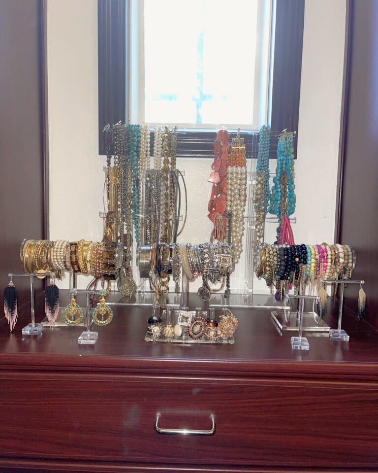 Styled jewelry display on a dark wood dresser in a walk-in closet, with necklaces, bracelets and earrings on clear acrylic stands, organized by Simply Luxe, Orange County