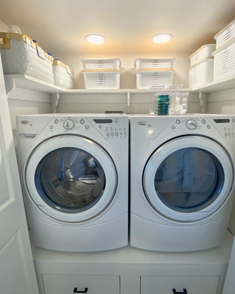 Laundry closet after organizing, with matching white bins on the shelf and clear canisters for detergent pods above the washer and dryer, organized by Simply Luxe, Lake Arrowhead