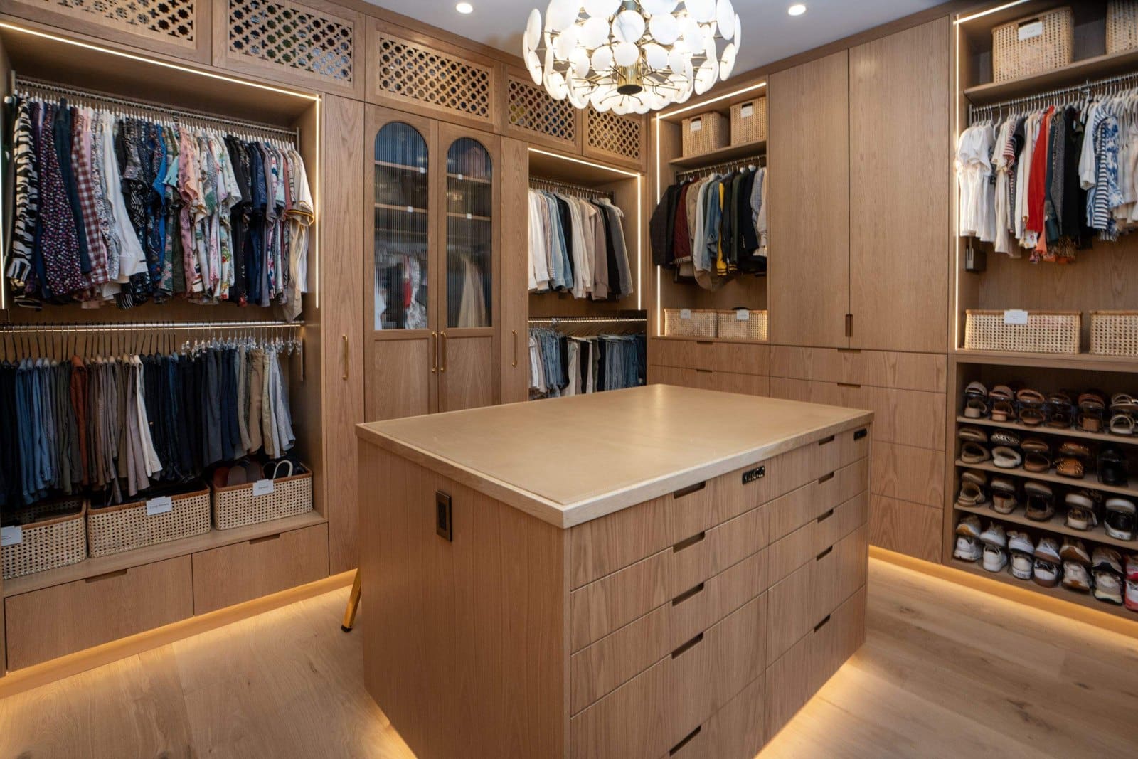 Custom warm-wood walk-in closet organized by Simply Luxe: color-sorted hanging clothes, labeled woven baskets, center island and shoe wall