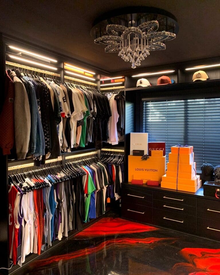 Men's closet after Simply Luxe organizing, with black hanging bays, lit drawers, Louis Vuitton trunks and a crystal chandelier above a glossy red and black floor, Costa Mesa
