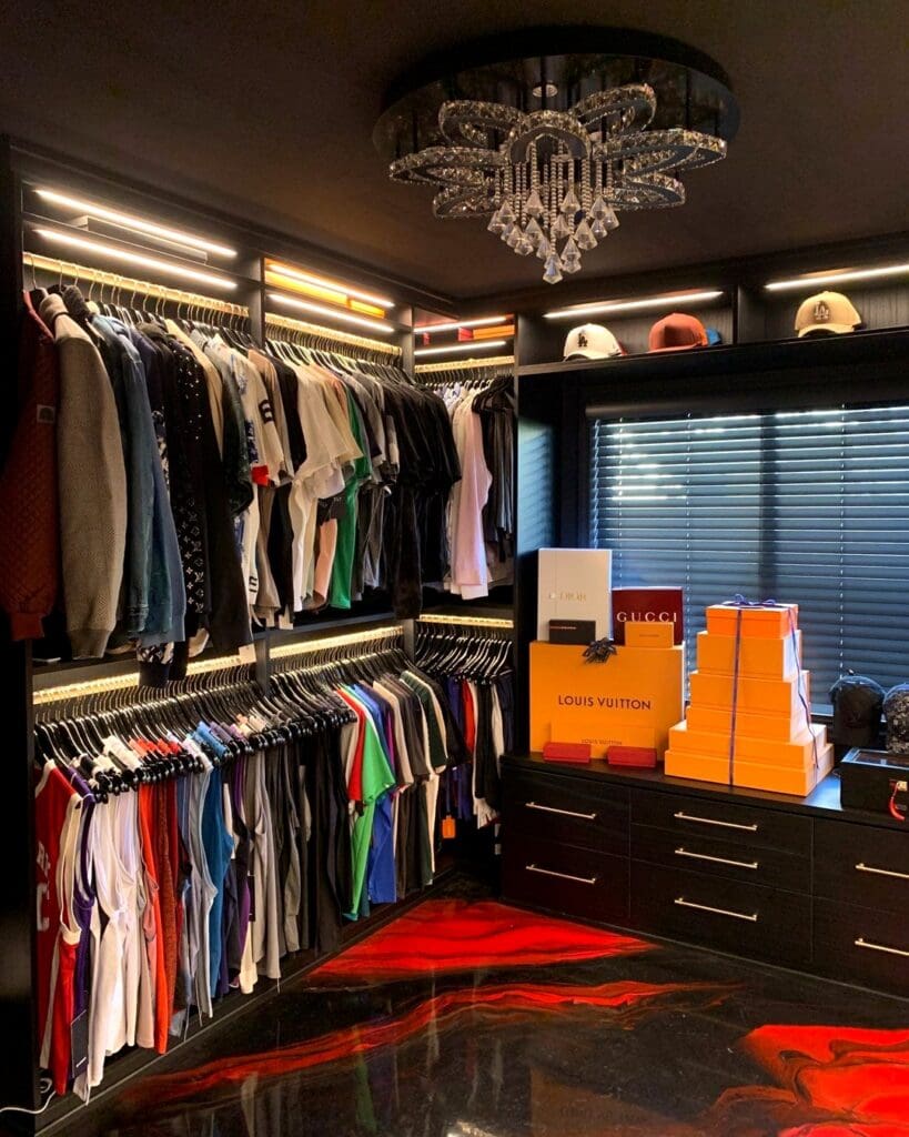 Men's closet after Simply Luxe organizing, with black hanging bays, lit drawers, Louis Vuitton trunks and a crystal chandelier above a glossy red and black floor, Costa Mesa