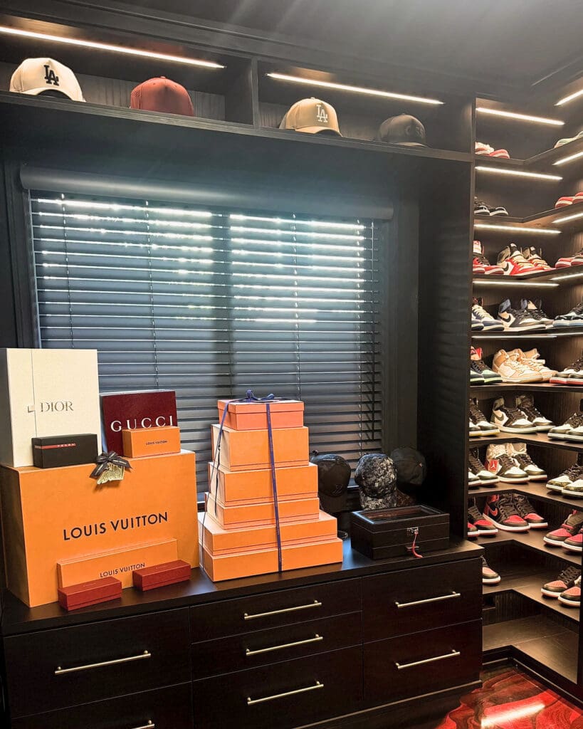 Same men's closet after Simply Luxe organizing, with sneakers displayed on lit shelves, caps lined up above the window and designer boxes styled on the dresser, Costa Mesa