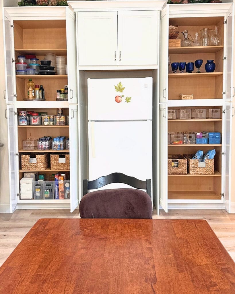 Same kitchen built ins after the Simply Luxe unpack, shelves filled with glassware, pantry baskets and dry goods around the fridge, San Diego