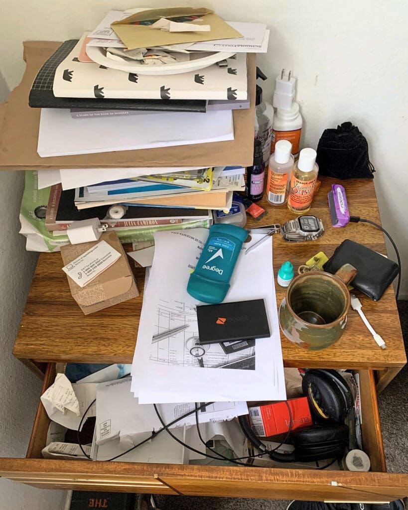 Bedside nightstand before organizing, buried under stacked papers, books and toiletries with an overflowing drawer, Costa Mesa home