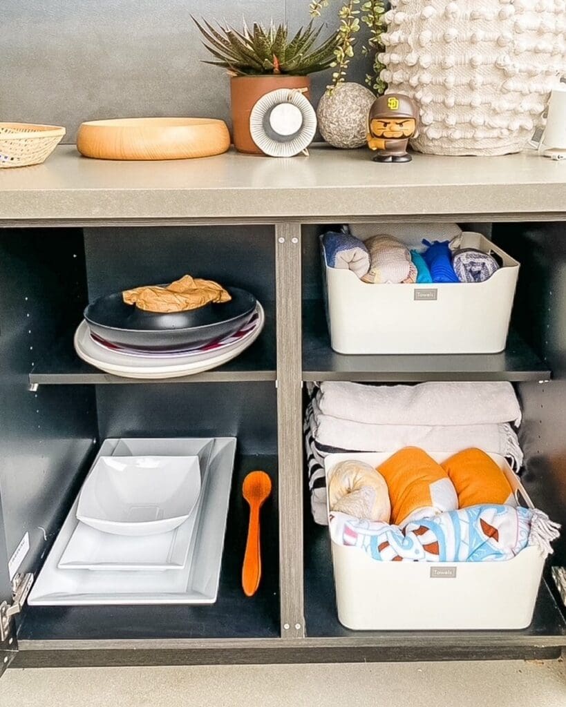 Same outdoor kitchen BBQ cabinet after organizing, with serving platters, plates and labeled towel bins, Del Mar