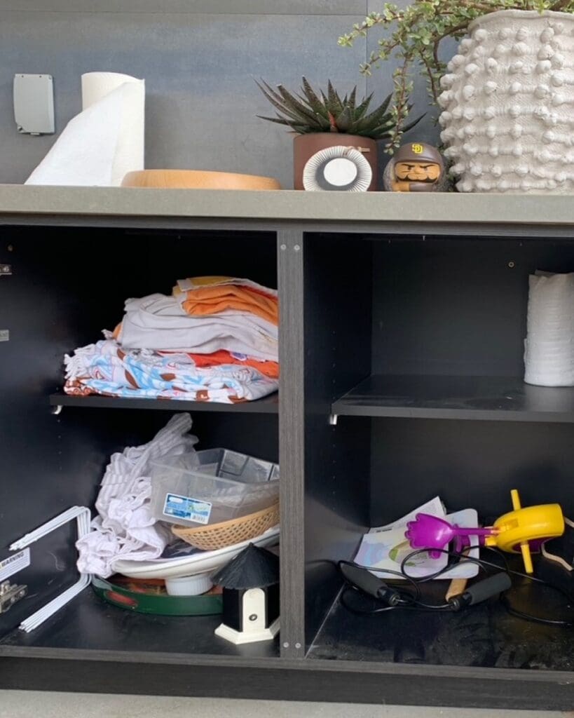 Outdoor kitchen BBQ cabinet before organizing, with towels, toys and loose items piled on the shelves, Del Mar