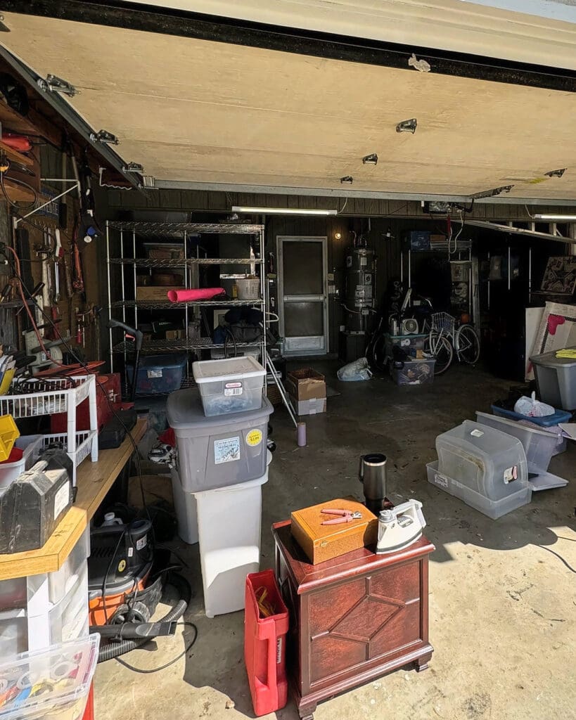 Two car garage before organizing, with stacked bins, furniture, tools and boxes scattered across the floor, Fountain Valley home